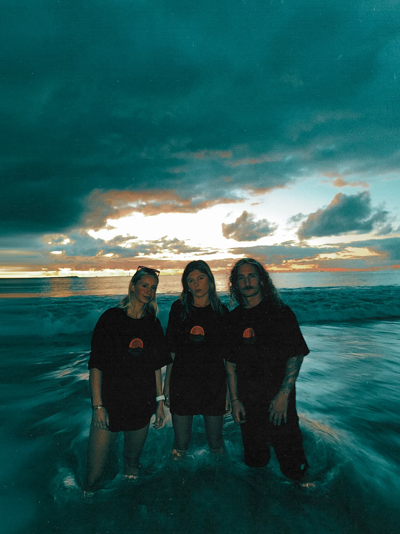 Three of the collective standing in the shallows at last light, all in the Coastline Social Club tee.